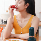 A woman in a yellow ribbed tank top holds a red fruit near her face while sitting at a table with a dark teal handheld cleaning device in the foreground. 