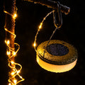 A hanging solar-powered string light with a warm white glowing globe and a dark solar panel top, shown against a black background.