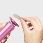 Hand holding a pink container with a small white object, against a light gray background.