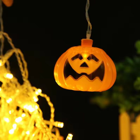 An illuminated orange jack-o'-lantern hanging light with a carved face, shown glowing against a dark night background with warm string lights blurred in the foreground.
