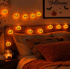 Warm white string lights shaped like glowing jack-o'-lanterns hanging above a bed in a cozy bedroom, with Halloween-themed bedding, a framed bat print, and a candle on the nightstand.