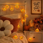 Warm white LED pumpkin string lights with glowing jack-o'-lantern faces draped over a bed in a cozy bedroom, with a nightstand, framed 'Trick or Treat' sign, and autumn decor in the background.