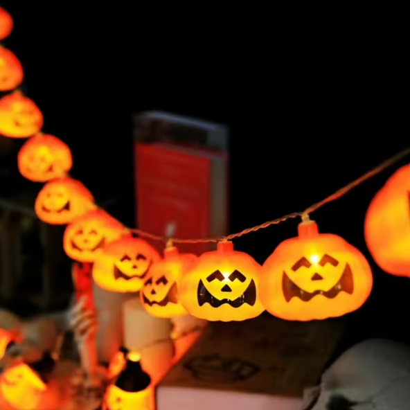 A string of glowing orange jack-o'-lantern string lights hanging against a dark background, with several pumpkins illuminated in the foreground and more visible along the cord.